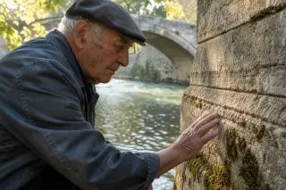 Elderly man in work clothes quietly examining mossy stonework at the base of an old bridge, river and trees softly lit in the background.