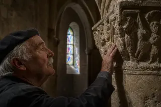 Elderly man quietly touching carved stone capital inside a Romanesque chapel, with Gothic arches blurred in background and soft colored light from a stained-glass window.