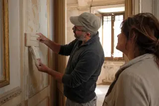 Artisan and homeowner examining mouldings in a sunlit hallway of an old Varois mansion, with dust motes visible, atmosphere subdued and contemplative.