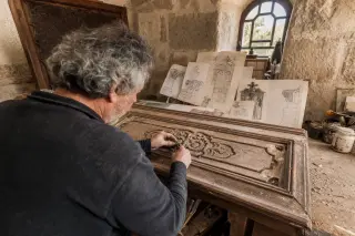 Artisan restoring an old carved wooden door panel in a stone-walled workshop, highlighted by soft natural light and surrounded by sketches and wood shavings.