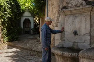Elderly man touches an old stone fountain in a shaded village alley, with a small oratory half-hidden by greenery in the background, soft Mediterranean morning light.
