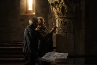 Elderly artisans study stone carvings in a softly lit Romanesque church interior, surrounded by ancient architectural details and warm, golden light.
