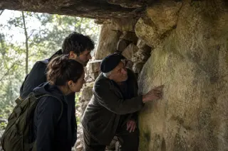 An elderly guide explains historical rock engravings to two young hikers inside a rustic stone shelter in a Var forest, with soft natural light and a warm, authentic feel.