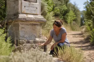 A local woman tends to a weathered stone oratory along a rural Var footpath, surrounded by wild Mediterranean plants in soft, natural light.