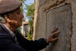 Elderly artisan's hands brushing dust from an ancient limestone block in a softly lit Var workshop, surrounded by restoration tools.