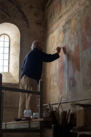 Elderly art restorer carefully working on a medieval fresco in a softly lit chapel, with natural Mediterranean light highlighting details of the ancient painting and calm, focused ambiance.