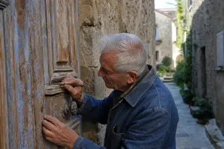 Elderly craftsman carefully restoring an old wooden door in a sunlit narrow Var village alley, surrounded by textured stone walls and soft Mediterranean light.