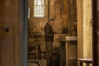 Elderly man polishing a candlestick inside an old stone chapel, sunlight highlighting aged frescoes and walls, evoking heritage and quiet tradition.