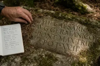 Hand brushing moss and pine needles from a Roman-inscribed stone along a wooded Var trail, with field notebook visible.